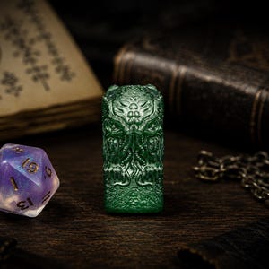 May include: A translucent green rectangular resin object featuring a detailed relief carving of a tentacled creature with multiple eyes. Beside it sits a twenty-sided die with a purple and white marbled finish and gold-colored numbers.