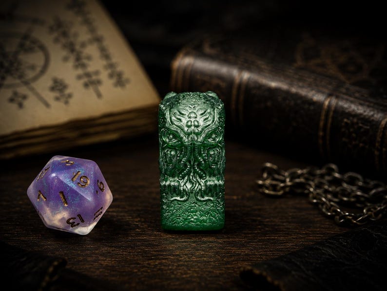 May include: A translucent green rectangular resin object featuring a detailed relief carving of a tentacled creature with multiple eyes. Beside it sits a twenty-sided die with a purple and white marbled finish and gold-colored numbers.