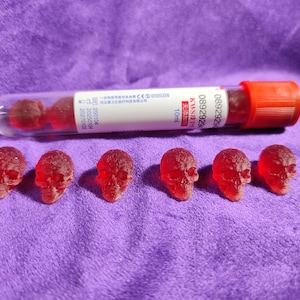 May include: A clear test tube with a red cap and a label, containing several red, round objects. Six small, red skull-shaped sweets are arranged below the tube on a purple surface. The tube has text on it.