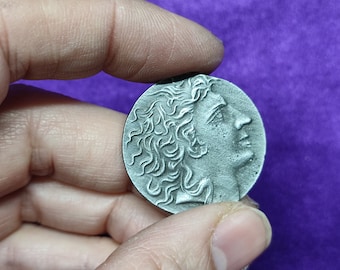 May include: A silver-colored coin featuring a profile of a person with curly hair. The coin has a detailed relief design and is held against a purple background.