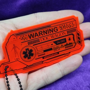 Puede incluir: Llavero de plástico naranja con una cadena negra. El llavero tiene un letrero de advertencia con el texto "WARNING DOC WAGON 24/365" y un símbolo médico.