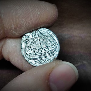 Może przedstawiać: Srebrna moneta z szczeg&oacute;łowym grawerunkiem statku wiking&oacute;w z masztem i żaglem. Moneta trzymana między palcami, ukazując jej skomplikowany wz&oacute;r i historyczny motyw. Powierzchnia monety ma lekko zużyty wygląd. Około 2.5 cm średnicy.