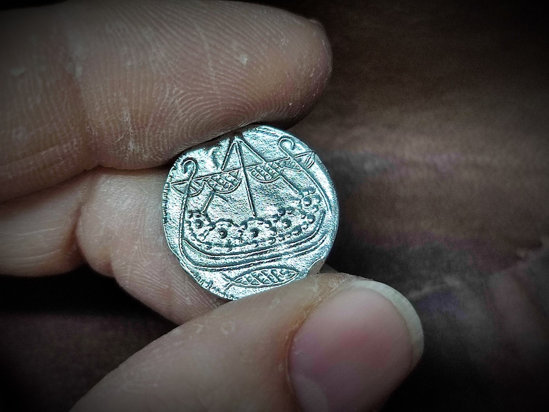 Może przedstawiać: Srebrna moneta z szczeg&oacute;łowym grawerunkiem statku wiking&oacute;w z masztem i żaglem. Moneta trzymana między palcami, ukazując jej skomplikowany wz&oacute;r i historyczny motyw. Powierzchnia monety ma lekko zużyty wygląd. Około 2.5 cm średnicy.