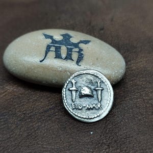 May include: A silver coin with a helmet and two swords on it. The coin has the words "Fid-Mar" inscribed on it.
