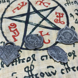 Puede incluir: Cuatro amuletos de metal plateado con diseños intrincados, que incluyen un pentagrama, un círculo con un nudo, un triángulo con un círculo y una luna creciente. Los amuletos están sobre un fondo de pergamino con marcas de tinta roja.