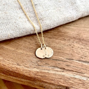 May include: A gold necklace featuring two small, circular pendants with engraved text. The delicate chain hangs against a wooden surface, with a linen cloth in the background. This necklace is a piece of jewellery.