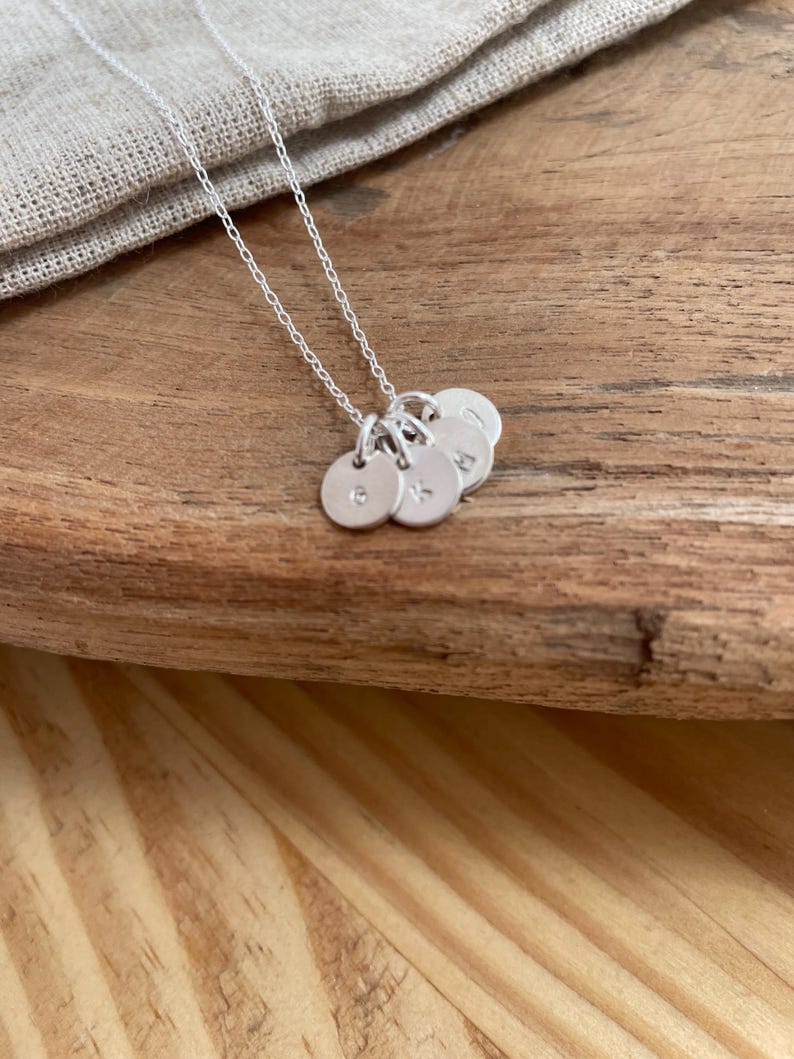 May include: A silver necklace with multiple small, round pendants, each bearing a single initial. The pendants are suspended from a delicate chain. The necklace rests on a wooden surface, with a neutral-colored fabric in the background.