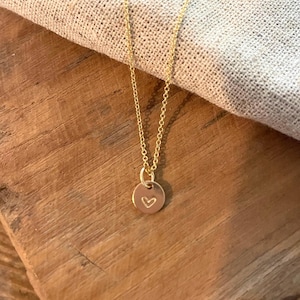 May include: A delicate gold-colored necklace with a small, round pendant. The pendant features a heart-shaped design. The necklace is displayed on a wooden surface with a neutral-colored fabric in the background.