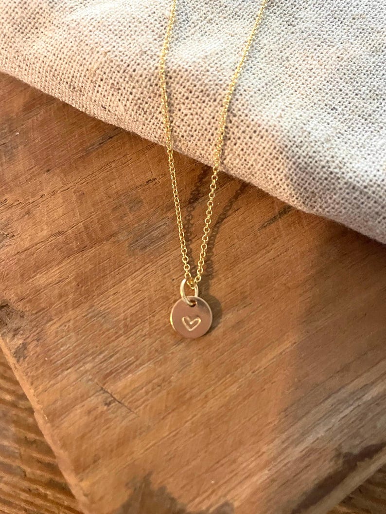 May include: A delicate gold-colored necklace with a small, round pendant. The pendant features a heart-shaped design. The necklace is displayed on a wooden surface with a neutral-colored fabric in the background.