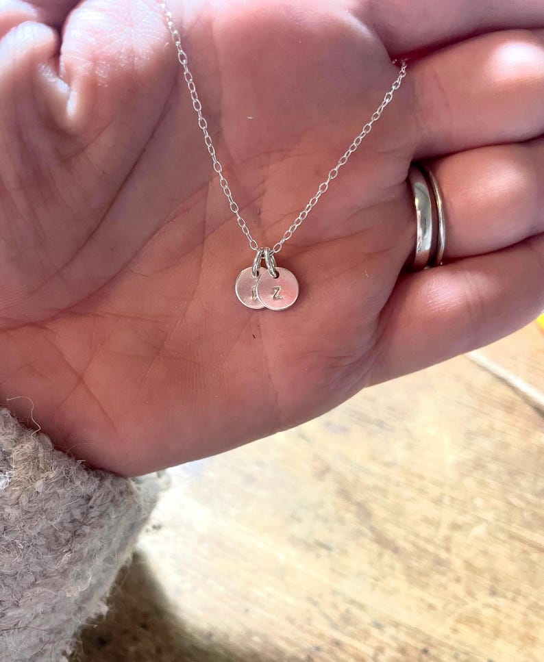 May include: A delicate silver necklace featuring two small, round pendants. Each pendant has a single letter engraved on it. The necklace is displayed on a hand, with a ring visible in the background. The chain is fine and the pendants are close together.