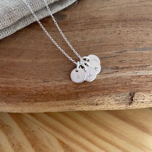 May include: A silver necklace with three small, round pendants. Each pendant has a single letter engraved on it. The necklace chain is delicate and the pendants are hanging on a wooden surface.