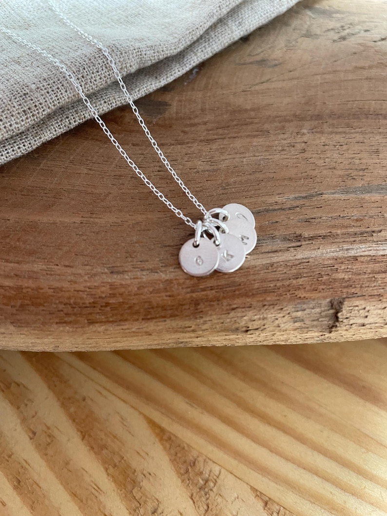 May include: A silver necklace with three small, round pendants. Each pendant has a single letter engraved on it. The necklace chain is delicate and the pendants are hanging on a wooden surface.
