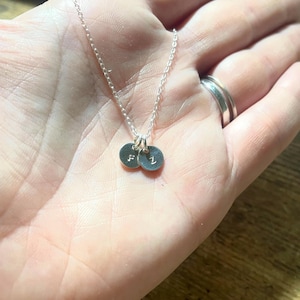 May include: A delicate silver necklace featuring two small, round pendants. Each pendant has a single initial engraved on it. The necklace is displayed on a hand, showcasing the fine chain and the personalized charms.