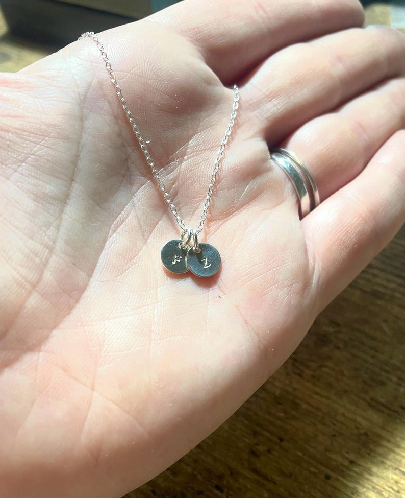 May include: A delicate silver necklace featuring two small, round pendants. Each pendant has a single initial engraved on it. The necklace is displayed on a hand, showcasing the fine chain and the personalized charms.