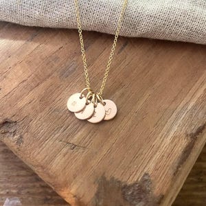 May include: A gold-colored necklace with a delicate chain and multiple small, round pendants. Each pendant has a single letter or symbol engraved on it. The necklace rests on a wooden surface.