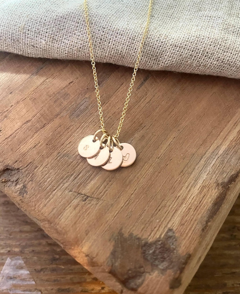 May include: A gold-colored necklace with a delicate chain and multiple small, round pendants. Each pendant has a single letter or symbol engraved on it. The necklace rests on a wooden surface.