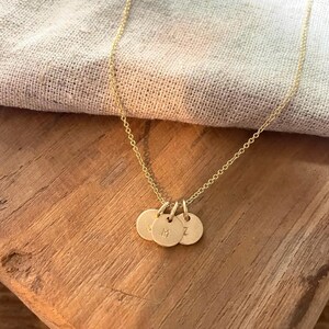 May include: A delicate gold-colored necklace featuring three small, round pendants. Each pendant has a single initial engraved on it. The necklace is displayed on a wooden surface with a soft, neutral-colored fabric in the background.