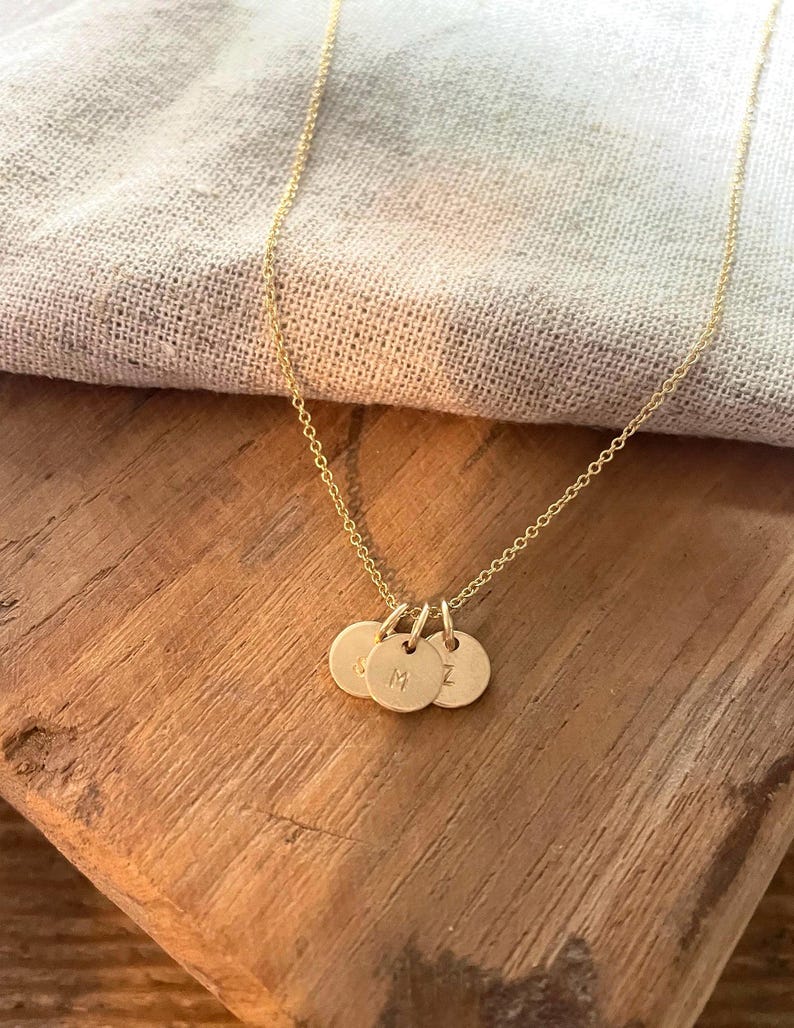 May include: A delicate gold-colored necklace featuring three small, round pendants. Each pendant has a single initial engraved on it. The necklace is displayed on a wooden surface with a soft, neutral-colored fabric in the background.