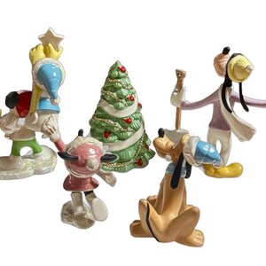 Może przedstawiać: Zestaw ceramicznych figurek bożonarodzeniowych przedstawiających postacie Disneya. Figurki obejmują Myszkę Miki, Minnie, Goofy'ego i Kaczora Donalda, a także udekorowaną choinkę. Postacie ubrane są w zimowe stroje.