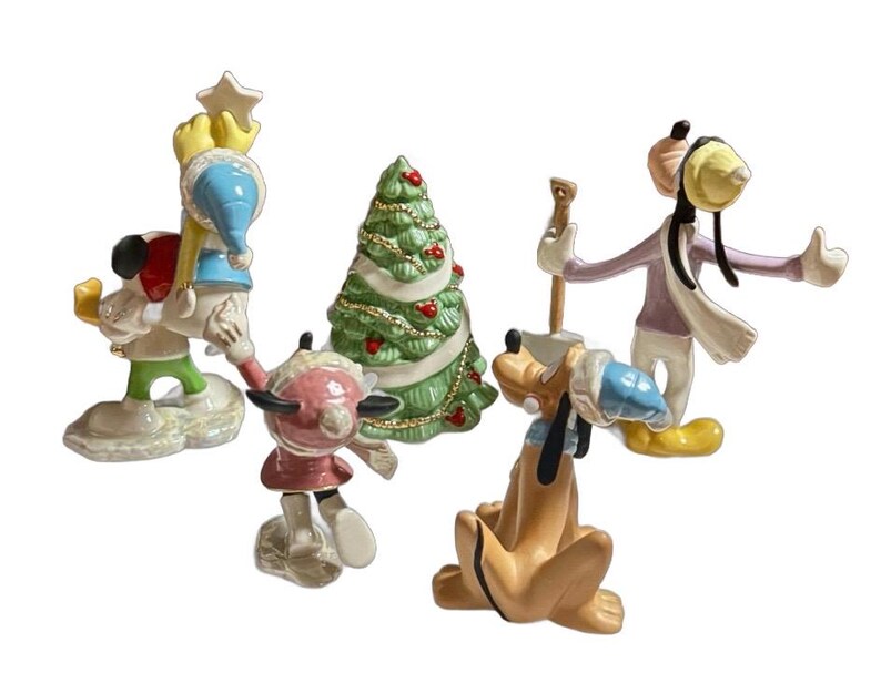 Może przedstawiać: Zestaw ceramicznych figurek bożonarodzeniowych przedstawiających postacie Disneya. Figurki obejmują Myszkę Miki, Minnie, Goofy'ego i Kaczora Donalda, a także udekorowaną choinkę. Postacie ubrane są w zimowe stroje.