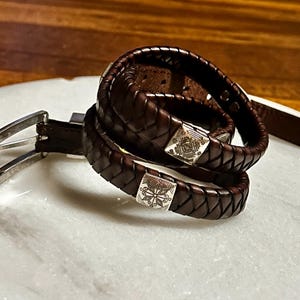 K&ouml;nnte beinhalten: Ein dunkelbrauner Lederg&uuml;rtel mit silberner Schnalle und ein geflochtenes Lederarmband mit quadratischen Silberakzenten. Der G&uuml;rtel und das Armband werden auf einer wei&szlig;en Marmoroberfl&auml;che pr&auml;sentiert.