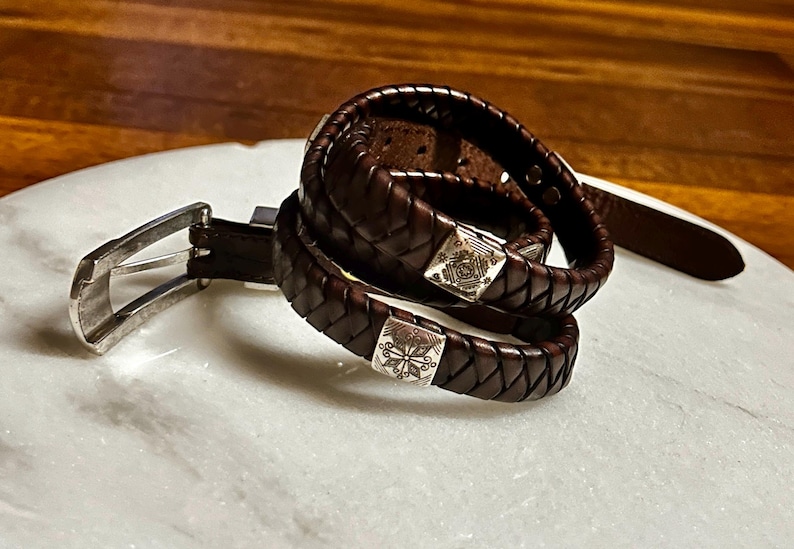 K&ouml;nnte beinhalten: Ein dunkelbrauner Lederg&uuml;rtel mit silberner Schnalle und ein geflochtenes Lederarmband mit quadratischen Silberakzenten. Der G&uuml;rtel und das Armband werden auf einer wei&szlig;en Marmoroberfl&auml;che pr&auml;sentiert.