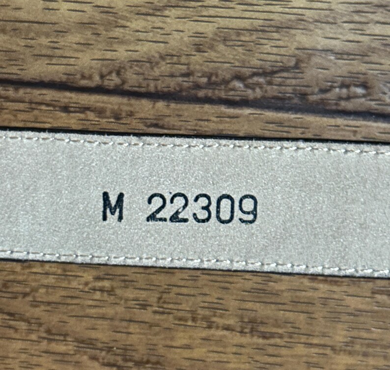 Puede incluir: Primer plano de una etiqueta de cuero beige con costuras negras y el texto "M 22309" impreso en ella.