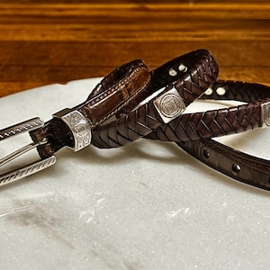 K&ouml;nnte beinhalten: Eine Sammlung brauner Lederg&uuml;rtel mit silbernen Schnallen und Akzenten. Ein G&uuml;rtel ist geflochten, ein anderer hat ein Krokodilmuster, und alle haben dekorative silberne Beschl&auml;ge. Die G&uuml;rtel werden auf einer wei&szlig;en Oberfl&auml;che pr&auml;sentiert.