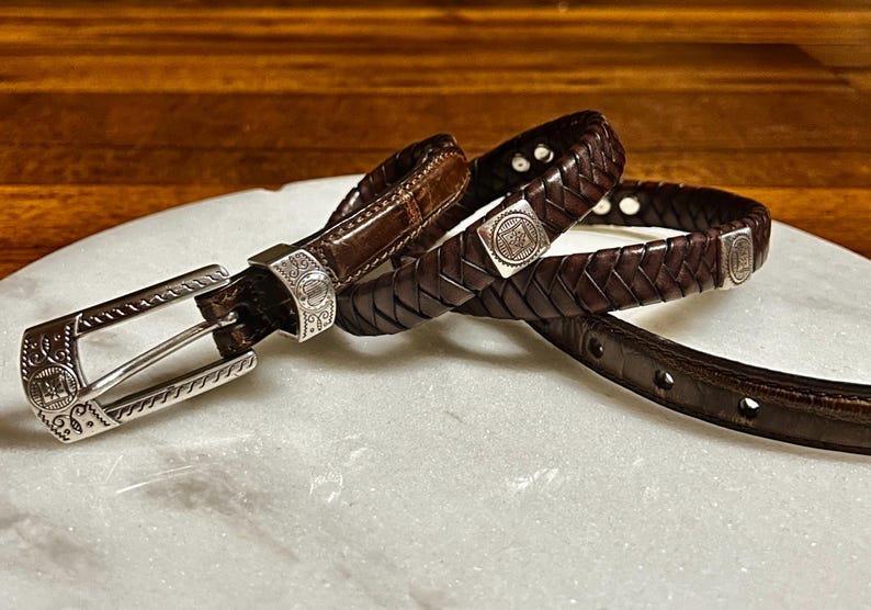 K&ouml;nnte beinhalten: Eine Sammlung brauner Lederg&uuml;rtel mit silbernen Schnallen und Akzenten. Ein G&uuml;rtel ist geflochten, ein anderer hat ein Krokodilmuster, und alle haben dekorative silberne Beschl&auml;ge. Die G&uuml;rtel werden auf einer wei&szlig;en Oberfl&auml;che pr&auml;sentiert.