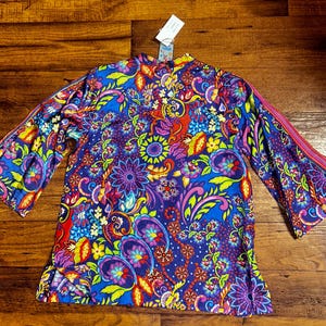 May include: A vibrant, long-sleeved top with a colorful floral and paisley pattern. The top features a mix of blue, purple, red, yellow, and green hues. The sleeves have a flared design with a pink stripe detail. A tag is attached at the top.