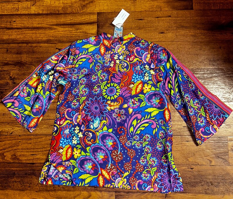 May include: A vibrant, long-sleeved top with a colorful floral and paisley pattern. The top features a mix of blue, purple, red, yellow, and green hues. The sleeves have a flared design with a pink stripe detail. A tag is attached at the top.