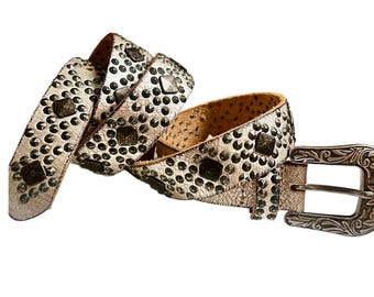 May include: A metallic silver belt featuring a textured surface adorned with rows of small round studs and larger square studs. The belt includes a silver-toned metal buckle with an intricate embossed floral pattern along its frame and prong.