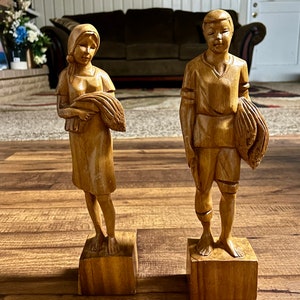 May include: Two wooden figurines of people, a woman and a man, standing on wooden bases. The woman is wearing a long dress and a headscarf, and she is holding a bundle of wheat. The man is wearing a short-sleeved shirt and pants, and he is holding a bundle of wheat.