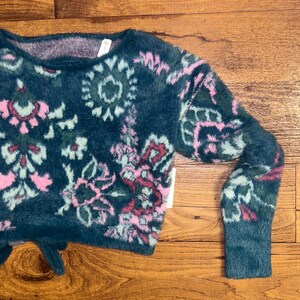 May include: A teal blue fuzzy sweater with a pink and green floral pattern. The sweater has a cropped length and long sleeves.