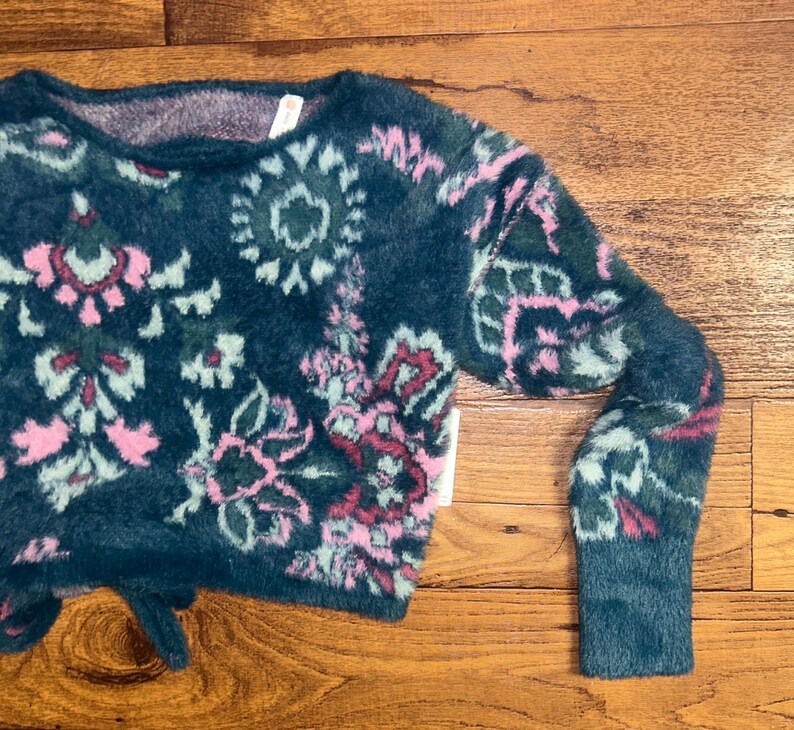 May include: A teal blue fuzzy sweater with a pink and green floral pattern. The sweater has a cropped length and long sleeves.