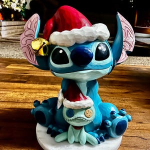 May include: A blue Stitch figurine wearing a red Santa hat with white trim and a gold bell. A smaller Stitch figure in a Santa hat sits in front. The figurine is on a white base and has a carved appearance.
