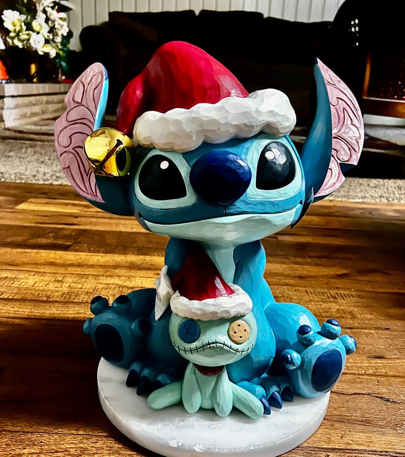 May include: A blue Stitch figurine wearing a red Santa hat with white trim and a gold bell. A smaller Stitch figure in a Santa hat sits in front. The figurine is on a white base and has a carved appearance.