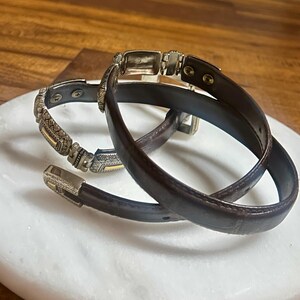 May include: Two brown leather belts with silver and gold-toned metal accents. One belt has a decorative buckle and a detailed pattern on the strap. The other belt is a simple design with a rectangular buckle. The belts are displayed on a white marble surface.