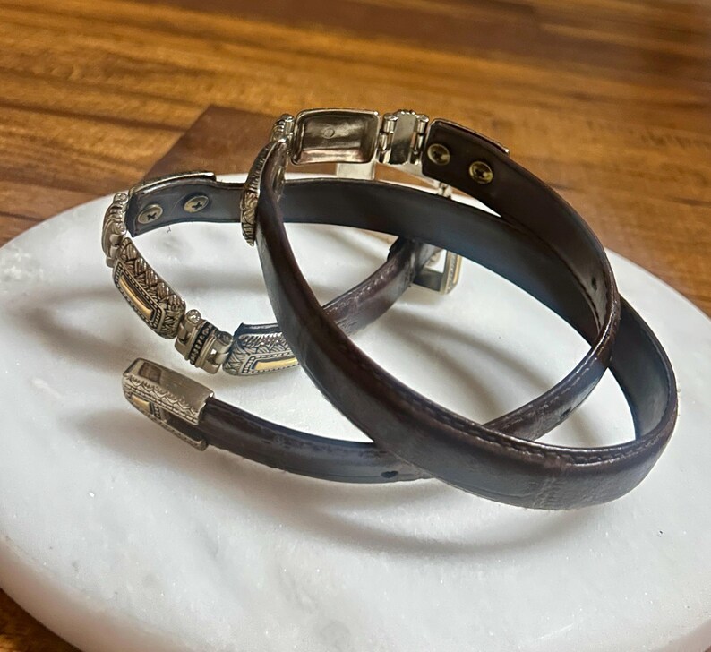 May include: Two brown leather belts with silver and gold-toned metal accents. One belt has a decorative buckle and a detailed pattern on the strap. The other belt is a simple design with a rectangular buckle. The belts are displayed on a white marble surface.