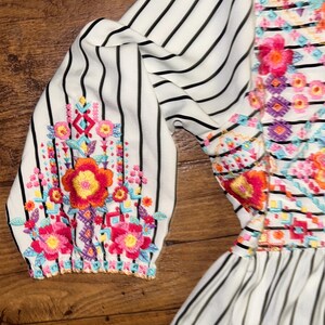 May include: White dress with black stripes and colourful floral embroidery. The dress has a ruffled hem and 3/4 sleeves.