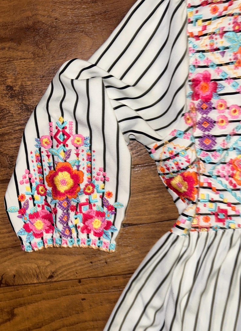 May include: White dress with black stripes and colourful floral embroidery. The dress has a ruffled hem and 3/4 sleeves.