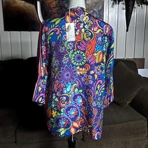 May include: A vibrant, long-sleeved kimono-style top with a colorful floral pattern. The design features a deep blue base with accents of red, orange, green, and purple. The garment is displayed on a mannequin, showcasing its flowing silhouette and intricate details.