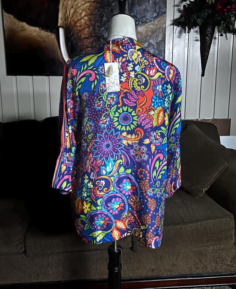 May include: A vibrant, long-sleeved kimono-style top with a colorful floral pattern. The design features a deep blue base with accents of red, orange, green, and purple. The garment is displayed on a mannequin, showcasing its flowing silhouette and intricate details.