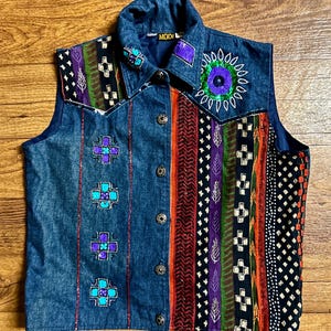May include: A denim vest with a colorful embroidered design. The vest has a collar and buttons. The embroidery features geometric patterns, flowers, and beads. The vest is a size medium.