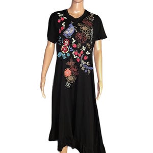 Puede incluir: Un vestido negro de largo midi con mangas cortas y cuello redondo. La parte delantera presenta bordados coloridos de un p&aacute;jaro azul, fresas rojas y varios patrones florales en tonos naranja, amarillo y azul, terminado con un dobladillo con volantes.