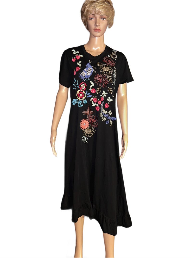 Puede incluir: Un vestido negro de largo midi con mangas cortas y cuello redondo. La parte delantera presenta bordados coloridos de un p&aacute;jaro azul, fresas rojas y varios patrones florales en tonos naranja, amarillo y azul, terminado con un dobladillo con volantes.