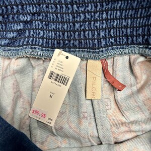 May include: Close-up of a denim garment with a smocked waistband. A white tag displays the size as M and the price. The garment has a patterned interior lining. The brand name "PILCRO" is visible on a small tag.