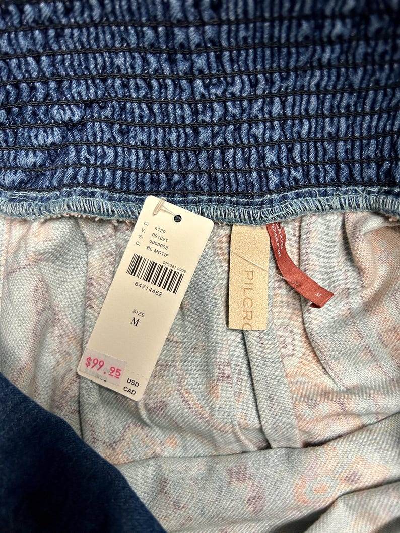 May include: Close-up of a denim garment with a smocked waistband. A white tag displays the size as M and the price. The garment has a patterned interior lining. The brand name "PILCRO" is visible on a small tag.