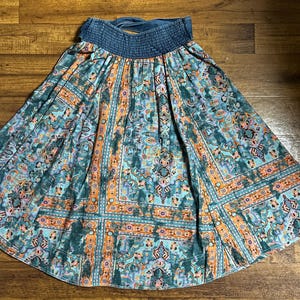 May include: A flowing skirt with a blue denim waistband and a vibrant, patterned design. The skirt features a mix of teal, orange, and purple hues in a geometric and floral print. The skirt is made of a lightweight fabric.