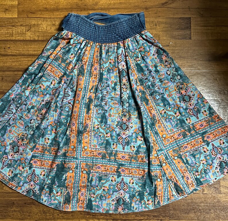 May include: A flowing skirt with a blue denim waistband and a vibrant, patterned design. The skirt features a mix of teal, orange, and purple hues in a geometric and floral print. The skirt is made of a lightweight fabric.